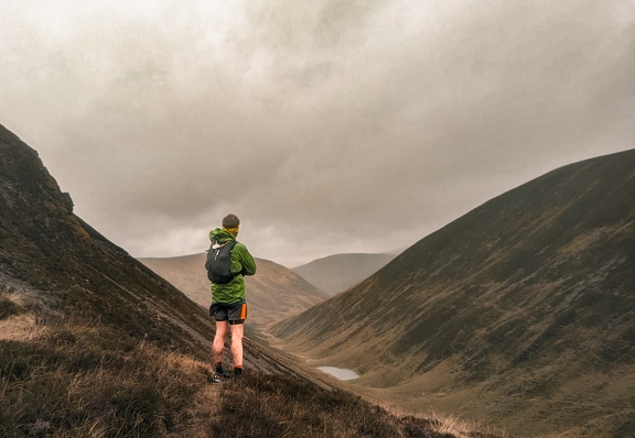 A photo of me, taken from behind, looking along a Glen. I'm wearing a green rain jacket and grey shorts. The hills are brown.